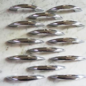 May include: A collection of polished, silver-toned cabinet pulls arranged on a light-colored surface. The pulls have a curved, ergonomic design, and are likely made of metal. The image showcases the hardware's reflective quality and sleek aesthetic.