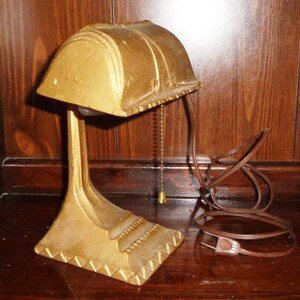 May include: A gold-colored desk lamp with a curved shade and a decorative base. The lamp has a cord and a pull chain.