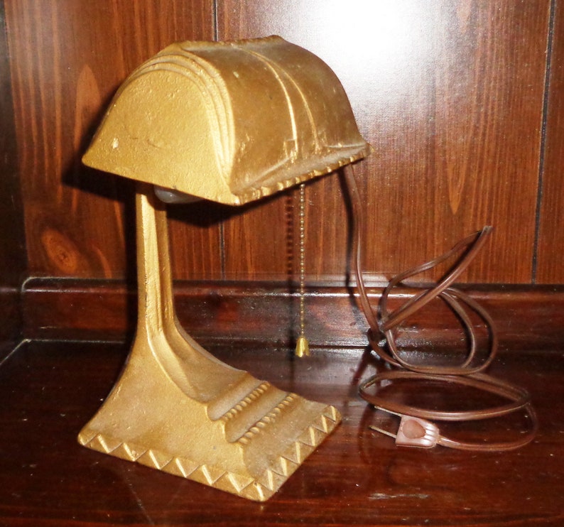 May include: A gold-colored desk lamp with a curved shade and a decorative base. The lamp has a cord and a pull chain.