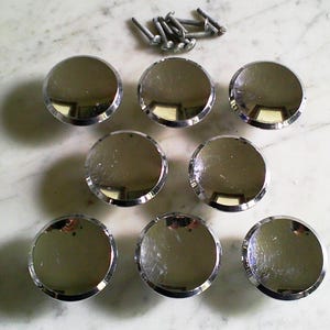 May include: A set of seven circular cabinet knobs with a polished chrome finish and a smooth, reflective surface. The knobs are arranged in three rows and are accompanied by seven matching metal mounting screws positioned at the top center.