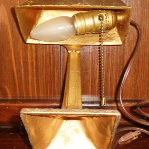 May include: A gold-colored desk lamp with a pull chain switch. The lamp has a simple design with a rectangular base and a single bulb.