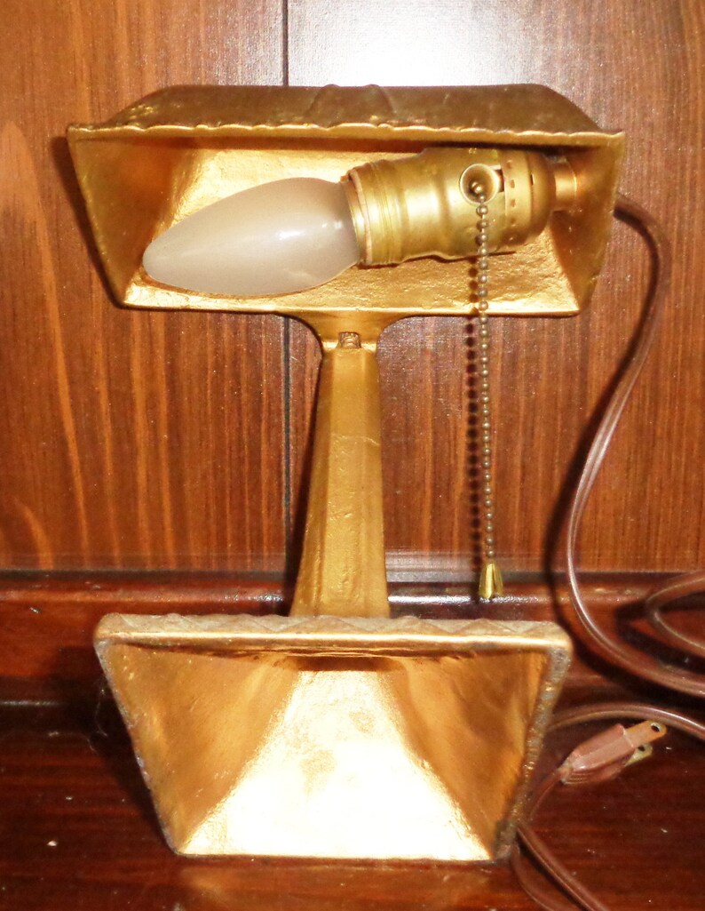 May include: A gold-colored desk lamp with a pull chain switch. The lamp has a simple design with a rectangular base and a single bulb.
