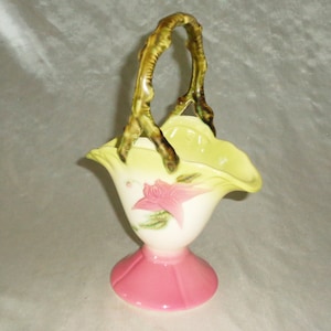 May include: A vintage ceramic basket with a green handle and a pink base. The basket is decorated with a white and green floral design.