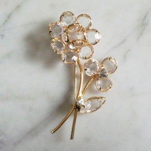 Puede incluir: Broche floral dorado con pétalos de cristal transparente facetado. El broche tiene un tallo y hojas, con una flor grande y un grupo de flores más pequeño. Los cristales brillan, sobre una superficie de mármol.