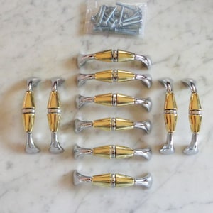 Puede incluir: Un juego de diez tiradores de armario con acabado dorado y plateado. Los tiradores tienen un diseño curvo con una sección central dorada y extremos plateados. Una pequeña bolsa de tornillos es visible en la parte superior de la imagen.