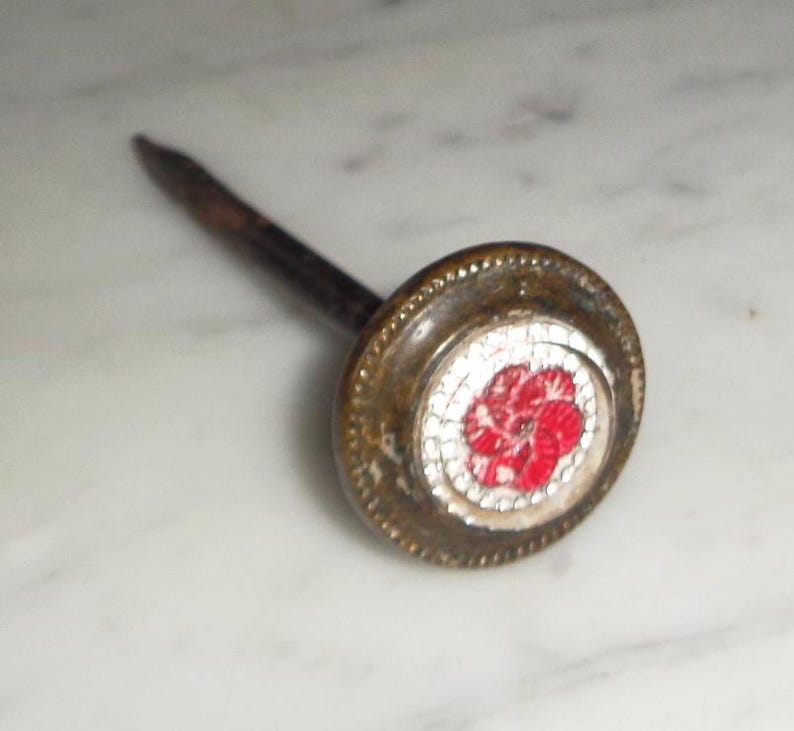 May include: A decorative nail with a round brass head featuring a red and white flower design.