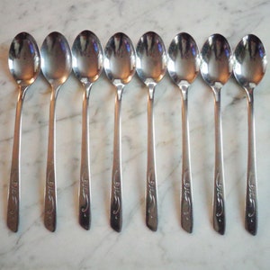 Puede incluir: Conjunto de ocho cucharas de plata con un diseño adornado. Las cucharas tienen un acabado pulido y están dispuestas en fila.