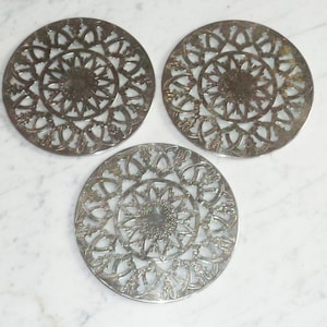 May include: Three silver metal coasters with a decorative, intricate, circular design. The coasters have a cut-out pattern that resembles a flower or star.