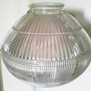 May include: A clear glass light fixture globe with a spherical shape. The upper section features vertical ribbed lines, while the lower section displays a textured grid pattern. The opening at the top is circular with a smooth, finished rim.