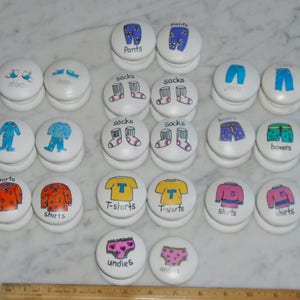 Pode incluir: Uma coleção de puxadores de gaveta brancos e redondos com ilustrações de roupas pintadas à mão. Cada puxador apresenta uma peça de roupa diferente, como camisas, calças, meias e roupa interior, com os nomes das roupas escritos abaixo.