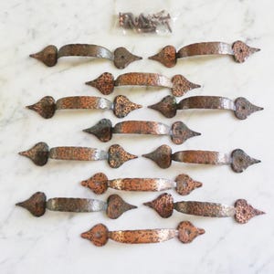 Könnte beinhalten: Zwölf antike Kupfer-Schranktürgriffe mit gehämmerter Oberfläche und spitzen Enden. Die Griffe sind in Reihen auf einer weißen Marmorfläche angeordnet.