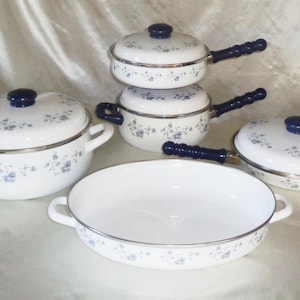 Op de afbeelding: Een witte emaille kookgerei set met blauwe bloempatronen. De set bevat een grote pot met handvatten, twee kleinere potten met handvatten, een koekenpan en deksels met donkerblauwe knoppen. De handvatten zijn donkerblauw.