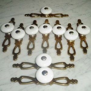 Könnte beinhalten: Satz von zehn weißen Keramikknöpfen mit silbernem Akzent und Messing-Schubladenschienen. Die Knöpfe und Schienen haben ein dekoratives Design.