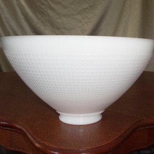 May include: A white, cone-shaped bowl with a textured, grid-like pattern. The bowl has a wide opening and a rounded base. The bowl is sitting on a dark brown, wooden table.
