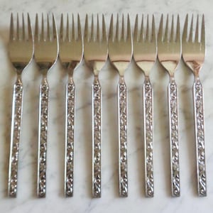 May include: A set of eight silver forks. Each fork has a tined head and a decorative handle with an embossed floral pattern. The forks are arranged in a row, reflecting light.