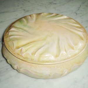 Puede incluir: Un plato de cerámica vintage con tapa, en una paleta de colores suaves y apagados de amarillo pálido, crema y toques de verde. El plato tiene un diseño texturizado en forma de hoja en la tapa y alrededor de la base, dándole un aspecto natural y orgánico. La forma general es redonda.