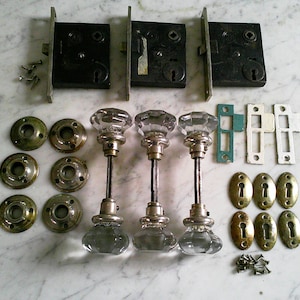 May include: An assortment of vintage door hardware, including three black mortise locks, six brass doorknob backplates, and three clear glass doorknobs with silver-toned stems. Also included are various strike plates and screws.