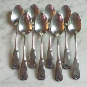 Puede incluir: Un juego de diez cucharas de plata con mangos adornados. Las cucharas están dispuestas en fila, con los mangos apuntando hacia el espectador.