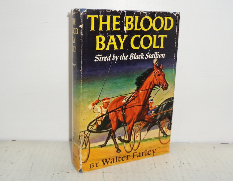 K&ouml;nnte beinhalten: Ein Vintage-Buchcover mit einem gelben und roten Titel "The Blood Bay Colt" und dem Untertitel "Sired by the Black Stallion". Das Cover zeigt ein Gem&auml;lde eines braunen Pferdes im Rennen mit einem Jockey.