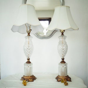 Puede incluir: Dos lámparas de mesa de cristal con pantallas blancas. Las lámparas tienen una base dorada y un diseño de cristal tallado.