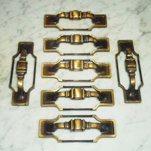 以下が含まれることがあります： 装飾的なデザインが施されたアンティークブラス製の引き出しノブ7個セット。各ノブは長方形で、中央部分が盛り上がっており、装飾的な縁取りがされています。