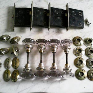 May include: An assortment of antique door hardware, including four glass doorknobs with silver-toned metal stems, several metal plates, and a black metal lock mechanism. Also included are various screws and keyhole covers.