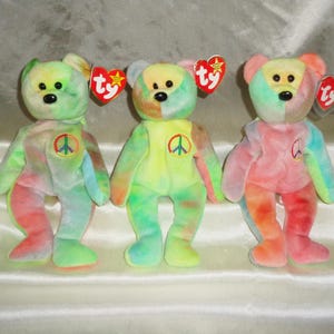 May include: Three tie-dye plush bears with peace symbols on their chests. Each bear has a different color scheme, including green, yellow, and pink. They have black button eyes and red heart-shaped tags.