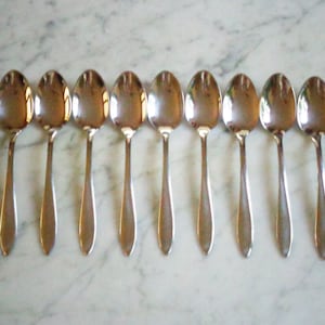 May include: Eight shiny, silver spoons arranged in a row. Each spoon has a rounded bowl and a long, slender handle. The spoons reflect the light, creating highlights and shadows.