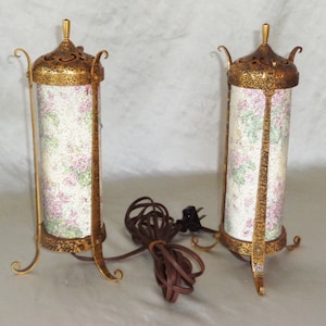 May include: Two gold-toned table lamps with floral patterned shades. The lamps have a decorative, ornate design and are on a white surface.