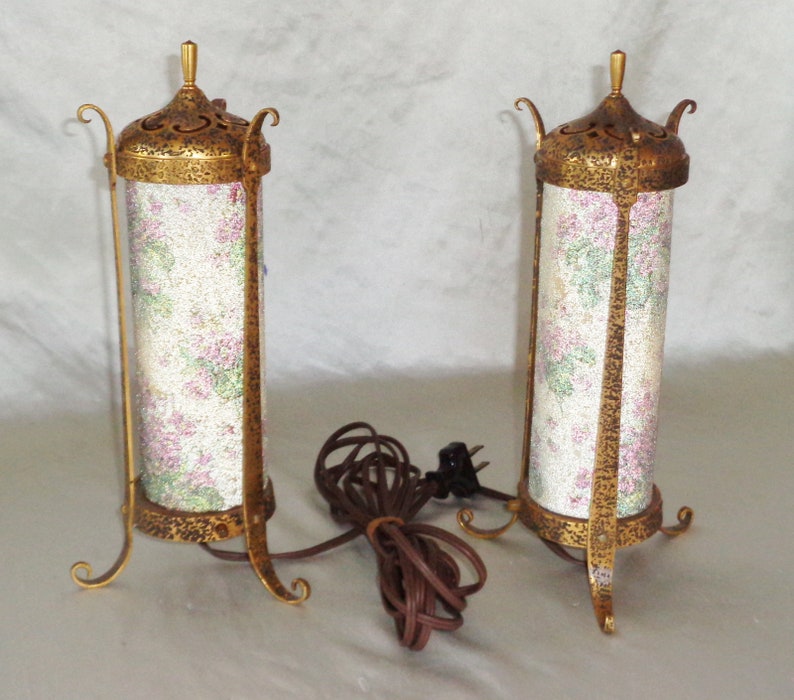 May include: Two gold-toned table lamps with floral patterned shades. The lamps have a decorative, ornate design and are on a white surface.