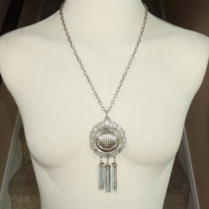 May include: A silver necklace with a large, round pendant featuring a geometric design and dangling tassels. The pendant is attached to a silver chain.