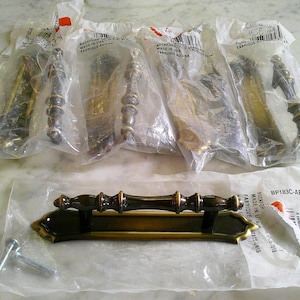 May include: A collection of five brass-toned metal cabinet pulls, each featuring a central handle with a decorative backplate. Four pulls are sealed in individual plastic packaging, while one is displayed outside its bag with two metal screws.