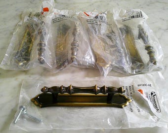 May include: A collection of five brass-toned metal cabinet pulls, each featuring a central handle with a decorative backplate. Four pulls are sealed in individual plastic packaging, while one is displayed outside its bag with two metal screws.