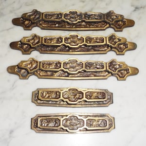 Puede incluir: Cinco tiradores de cajón de latón antiguo con un diseño ornamentado. Los tiradores son todos del mismo tamaño y forma, y tienen un acabado ligeramente texturizado.