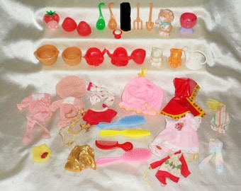 Può includere: Una collezione di accessori per bambole Strawberry Shortcake, tra cui vestiti, scarpe, un annaffiatoio, una pentola, una spazzola e un pettine. Gli accessori sono rosa, rosso, bianco e giallo.