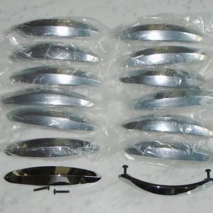 Könnte beinhalten: Ein Satz mit 10 silbernen Schrankgriffen mit einem geschwungenen, modernen Design. Die Griffe sind einzeln in durchsichtigen Plastiktüten verpackt. Außerdem gibt es einen einzelnen, größeren silbernen Griff mit einem geschwungenen Design.