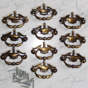 Puede incluir: Diez pomos de armario de latón antiguo con un diseño decorativo. Los pomos tienen un centro elevado y una forma curva. Los pomos se acompañan de un juego de tornillos.