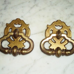 Puede incluir: Dos tiradores de puerta de armario de latón antiguo con diseños ornamentados. Los tiradores tienen un asa en forma de anillo y una parte superior decorativa.