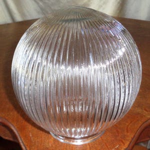 May include: A clear, spherical glass lampshade with vertical ribbed detailing. The lampshade has a rounded shape and a slightly flared base. The glass appears to be transparent, allowing light to pass through.