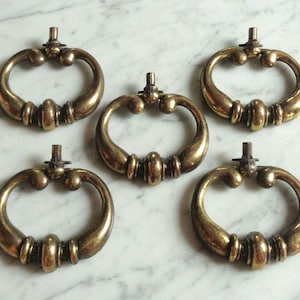 May include: Five vintage brass drawer pulls, each with a ring-shaped handle. The pulls have a central, cylindrical detail and a mounting post. The brass has a warm, aged patina, indicating a vintage or retro aesthetic.