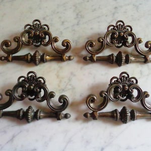 May include: Four antique bronze drawer pulls with ornate, scrollwork designs. Each pull features a central, detailed medallion and curved handles. The hardware is likely for furniture restoration or decorative use.