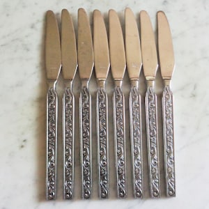 May include: A set of eight silver-colored knives. Each knife has a blade and a handle with an ornate floral design. The knives are arranged in a row on a light-colored surface.