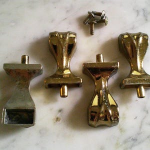 Op de afbeelding: Vier metalen meubelpoten met een klauwpootontwerp, voorzien van een gepolijste goudkleurige afwerking op drie stuks en een zilverkleurige afwerking op &eacute;&eacute;n stuk. De set bevat vier kleine metalen schroeven voor bevestiging, elk met een ronde kop.