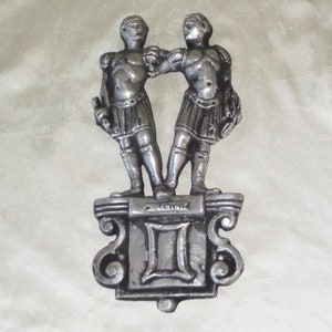 Puede incluir: Decoraci&oacute;n de pared de metal plateado con el signo del zod&iacute;aco G&eacute;minis. Dos figuras se paran sobre un pedestal con la palabra "G&eacute;minis" y el s&iacute;mbolo II.