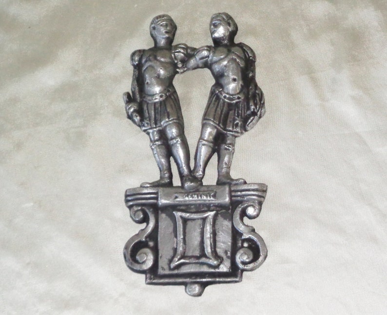 Puede incluir: Decoraci&oacute;n de pared de metal plateado con el signo del zod&iacute;aco G&eacute;minis. Dos figuras se paran sobre un pedestal con la palabra "G&eacute;minis" y el s&iacute;mbolo II.