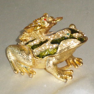 Pode incluir: Caixa de joias de sapo dourado com um sapo menor no topo. O sapo maior tem esmalte verde e preto com strass. O sapo menor tem olhos vermelhos.