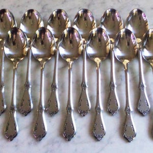 Puede incluir: Una colección de doce cucharas de plata dispuestas en fila. Cada cuchara tiene un cuenco redondeado y un mango largo y delgado con un diseño ornamental, similar a una flor, en la base. Las cucharas son reflectantes.