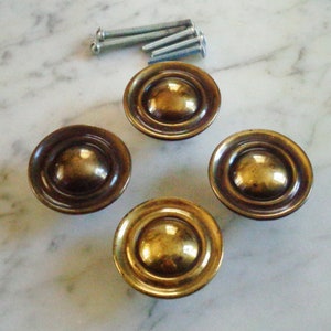 Może przedstawiać: Cztery złote, okrągłe gałki do szafki z wypukłym środkiem i koncentrycznymi okręgami. Gałki znajdują się na białym marmurowym blacie.