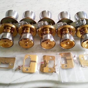 May include: A set of five vintage door knobs with matching hardware. The knobs have a two-tone design with a silver top and gold base. The set includes gold-colored strike plates and screws, all arranged on a marble surface.