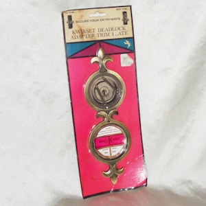 Puede incluir: Una placa de acabado de adaptador de pestillo Kwikset de color dorado empaquetada en un fondo rojo y azul. El paquete dice "Secure Your Entryways" y "Kwikset Deadlock Adapter Trim Plate".